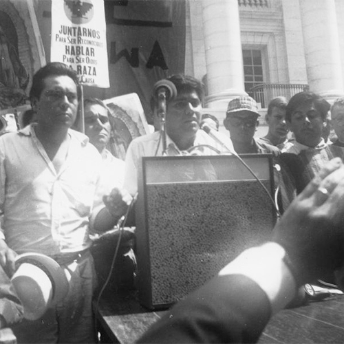 Jesus Salas addresses a rally at a podium in front of the Wisconsin State Capitol. Behind him are marchers who began the march from Wautoma and walked to Madison on Highway 21. Marchers are holding images of Our Lady of Guadalupe, a National Farm Worker Association's banner, and signs with the message 'Juntarnos para ser reconocidos. Hablar para ser oidos. La raza tiene causa'/ 'Join us to recognized. Speak to be heard. The people have a cause.'            This photograph is a part of Wisconsin-native David Giffey's series 'Struggle for Justice,'' images from the migrant farm worker struggle including an independent oganizing effort in Wisconsin and the nationwide grape boycott movement started by Cesar Chavez of United Farm Workers during the 1960s and 1970s.            Salas se dirige al público            Jesús Salas le habla al público desde un podio enfrente del Capitolio del estado de Wisconsin. Detrás de él están unos manifestantes que marcharon desde Wautoma y caminaron hacia Madison en la carretera Highway 21. Unos manifestantes están sosteniendo imágenes de Nuestra Señora de Guadalupe, el estandarte de National Farm Workers Association (NFWA), y letreros que dicen, 'Juntarnos para ser reconocidos. Hablar para ser oídos. La raza tiene causa.' Esta fotografía es parte de la serie 'Lucha por la Justicia' tomada por David Giffey, originario de Wisconsin, las imágenes muestran la lucha de los trabajadores agrícolas emigrantes incluyendo un esfuerzo independiente organizado en Wisconsin y el movimiento nacional del boicot de uvas empezado por Cesar Chávez de la unión de campesinos o United Farm Workers durante los años 1960 y 1970.r